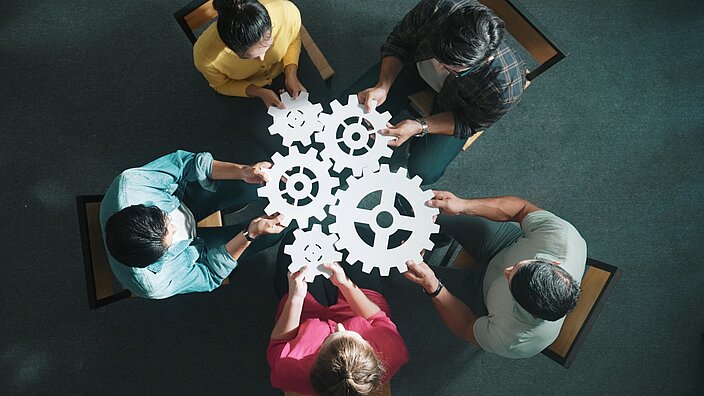 Draufsicht auf 5 Personen, die in ihren Händen weiße Zahnräder aneinander halten.