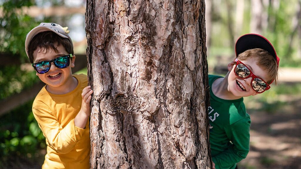 Zwei Kinder mit Sonnenbrillen hinter einem Baum