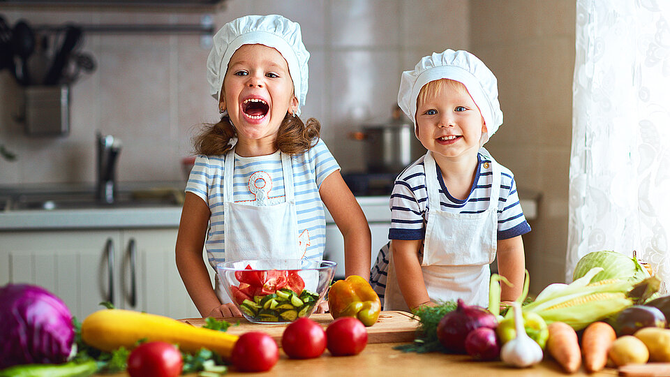 Zwei Kinder mit Kochmützen haben Spaß beim gesunden Kochen
